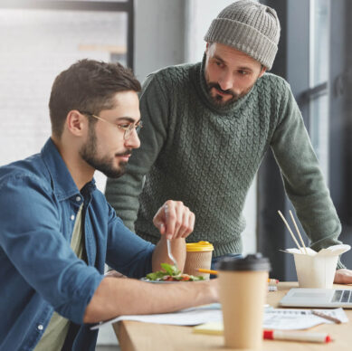Two men working together at a desk with laptops and coffee cups, one eating a salad while the other gives feedback