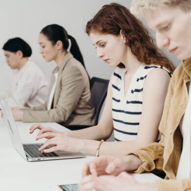 Focused coworkers sitting in a row at a desk, working on laptops and tablets in a modern office environment