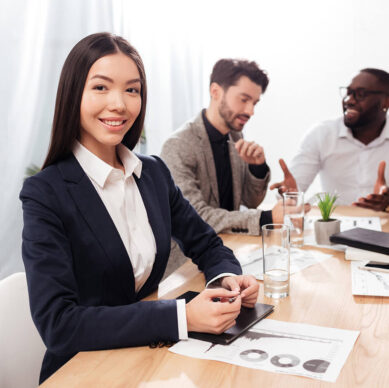 Business team having a meeting in a bright office; a confident woman in a suit smiles at the camera while two colleagues discuss work in the background
