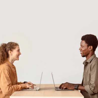 Two colleagues sitting across from each other at a desk, smiling and working on laptops in a minimal office setting