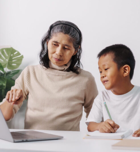 Learning online maths class on laptop computer pc, Asian grandso Asian grandson and grandmother people sitting together at desk doing homework at home.