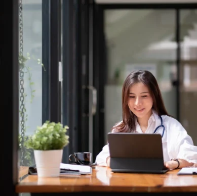 Smiling Asian female doctor in a hospital using a laptop while wearing a white coat and stethoscope