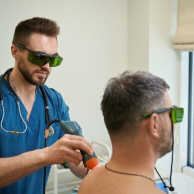 A male healthcare professional wearing blue scrubs and protective green glasses performs a laser therapy treatment on the upper back of a seated male patient, who is also wearing protective glasses, in a well-lit medical room near a window