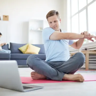 Man exercising at home while watching workout video on laptop, woman sitting on sofa in background